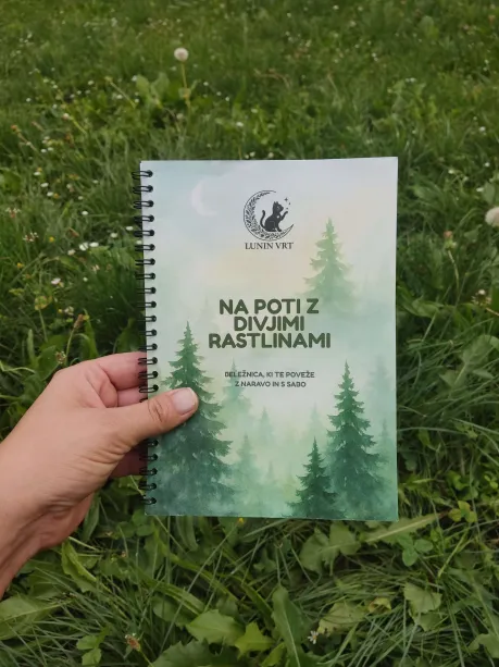 Oseba drži nabiralniško beležnico Na poti z divjimi rastlinami znamke Lunin vrt na travniku, obdana z naravo in travniškimi cvetovi.